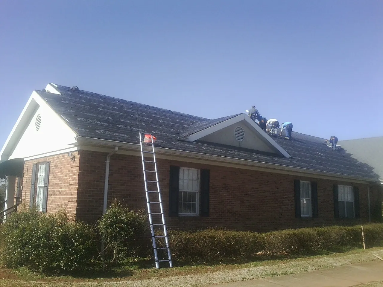 Crew tearing shingles off a brick ranch roof with a ladder set — Metal Roof Repair work by our crew in Huguenot