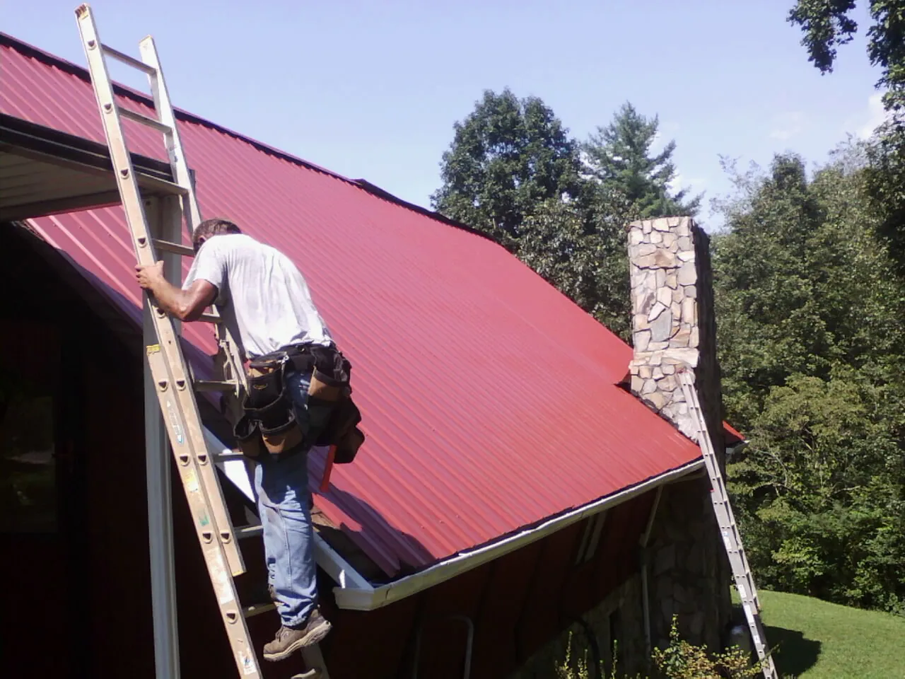 Roofer on a ladder beside a red metal roof and stone chimney — Soffit & Fascia Repair by Roofing Brothers in Huguenot